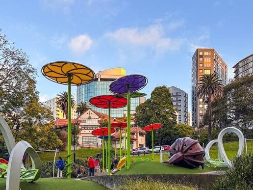 An inner-city playground with flower-shaped shade canopies and a snail sculpture, apartment towers behind it